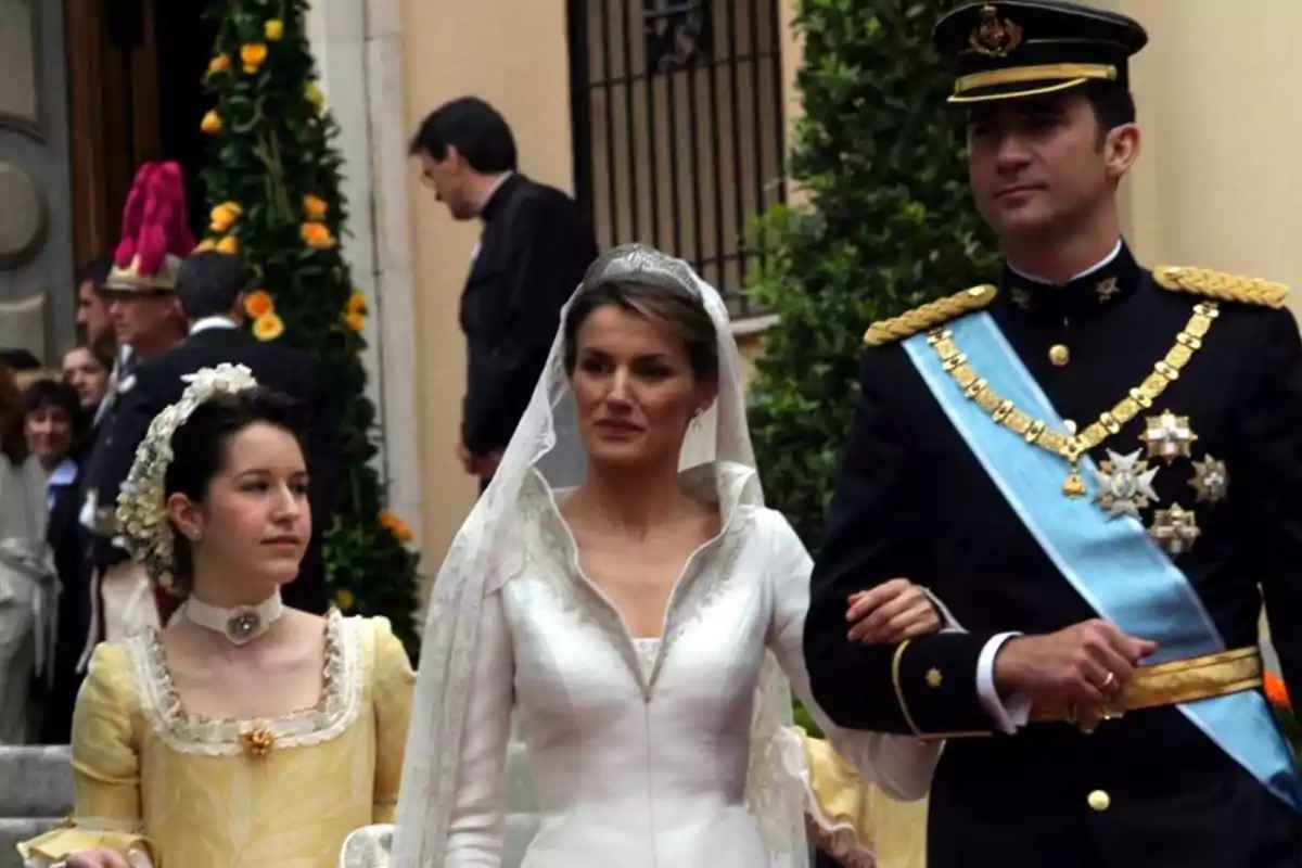 Una pareja vestida de manera formal camina junto a una joven en un evento al aire libre con flores y personas de fondo. Una pareja vestida de manera formal camina junto a una joven en un evento al aire libre con flores y personas de fondo.