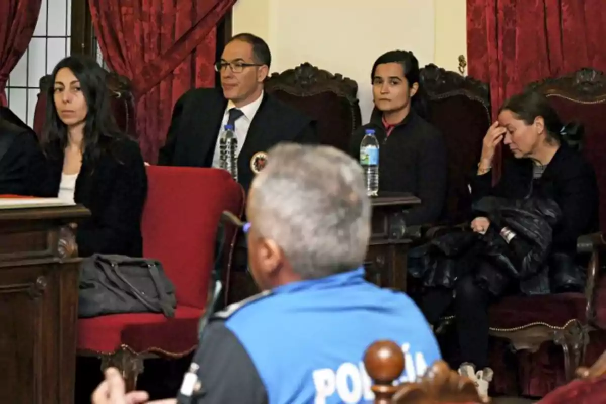 Un grupo de personas sentadas en una sala con cortinas rojas, algunas parecen estar en una audiencia o reunión formal.