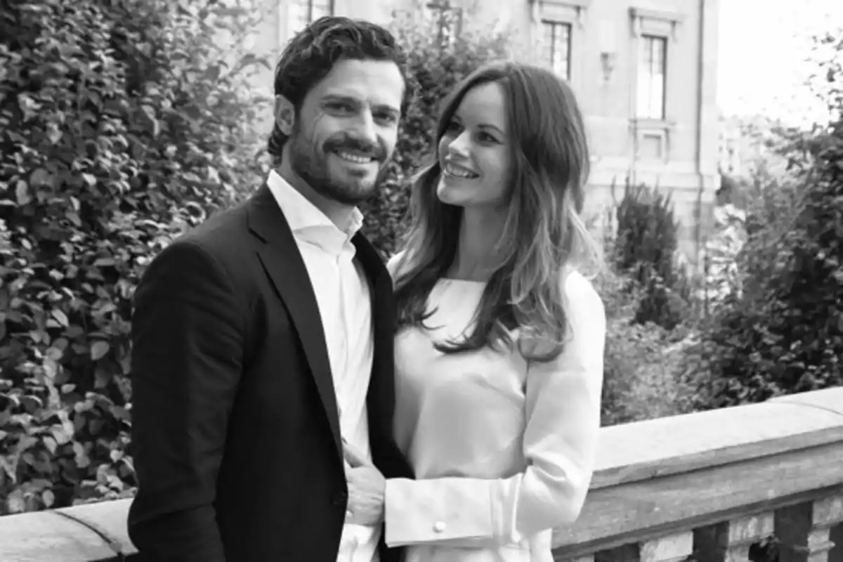 Pareja sonriente posando al aire libre junto a una barandilla con vegetación y un edificio de fondo en una imagen en blanco y negro