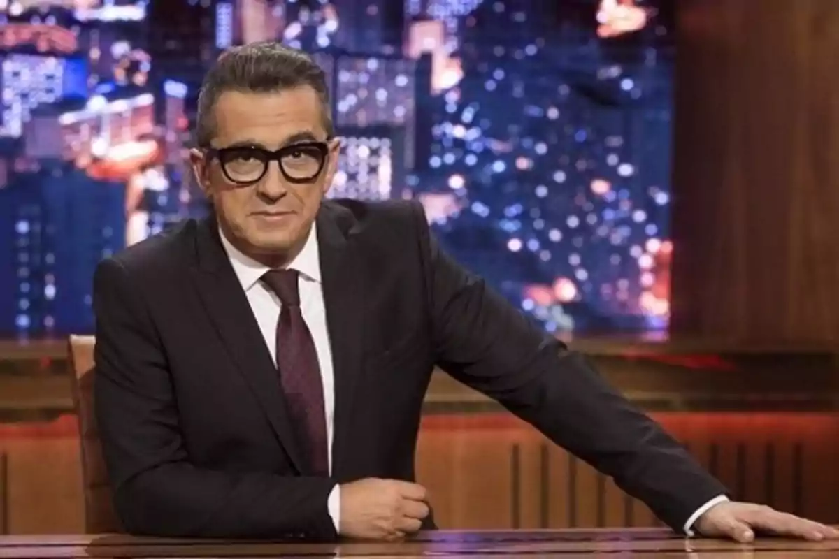 Un hombre con gafas y traje oscuro está sentado en un escritorio con un fondo de luces de ciudad. Un hombre con gafas y traje oscuro está sentado en un escritorio con un fondo de luces de ciudad.