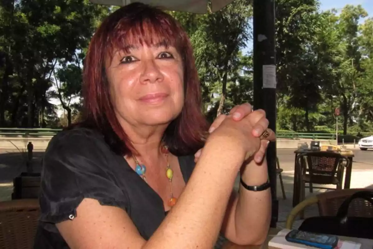 Mujer de cabello rojizo sentada en una terraza al aire libre con árboles de fondo y las manos entrelazadas sobre la mesa Mujer de cabello rojizo sentada en una terraza al aire libre con árboles de fondo y las manos entrelazadas sobre la mesa