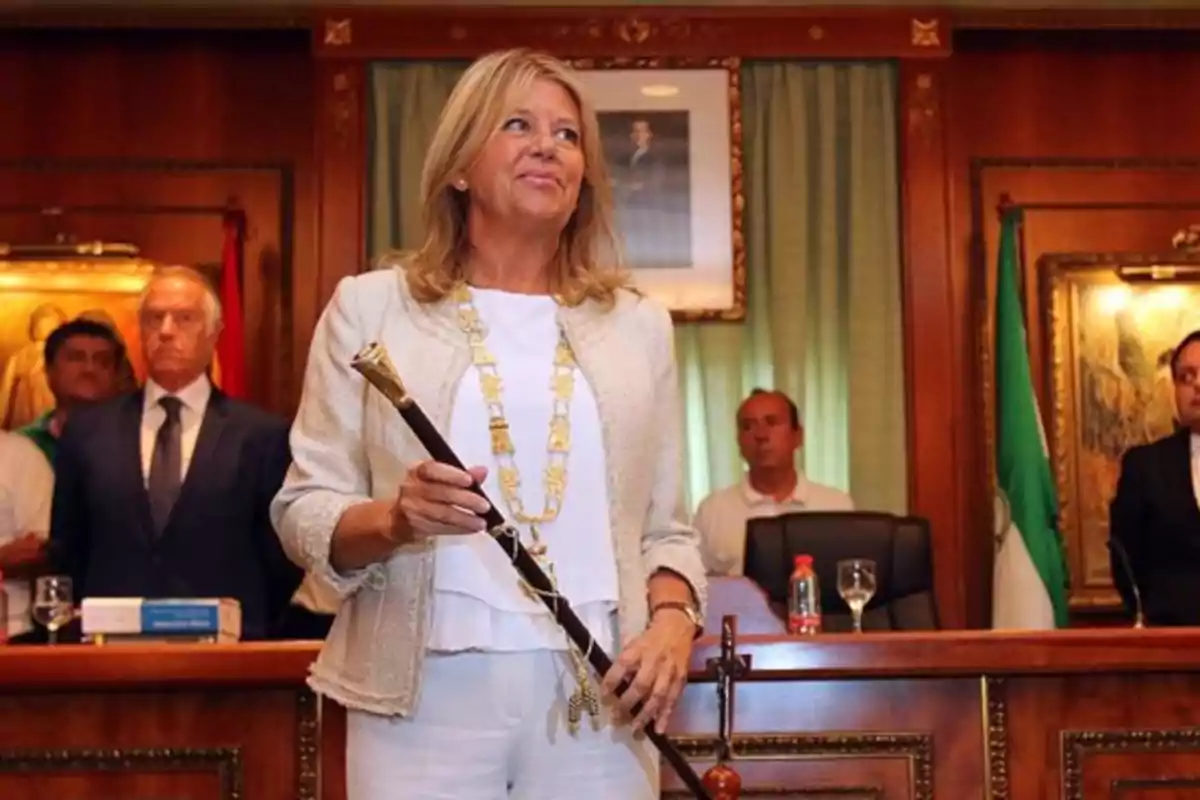 Mujer rubia vestida de blanco sosteniendo un bastón de mando en una sala de gobierno con varias personas al fondo Mujer rubia vestida de blanco sosteniendo un bastón de mando en una sala de gobierno con varias personas al fondo