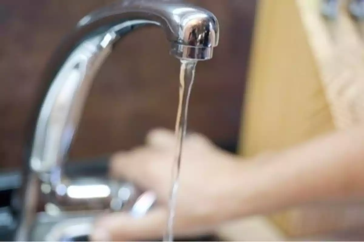 Un grifo de metal con agua fluyendo y una mano desenfocada en el fondo.