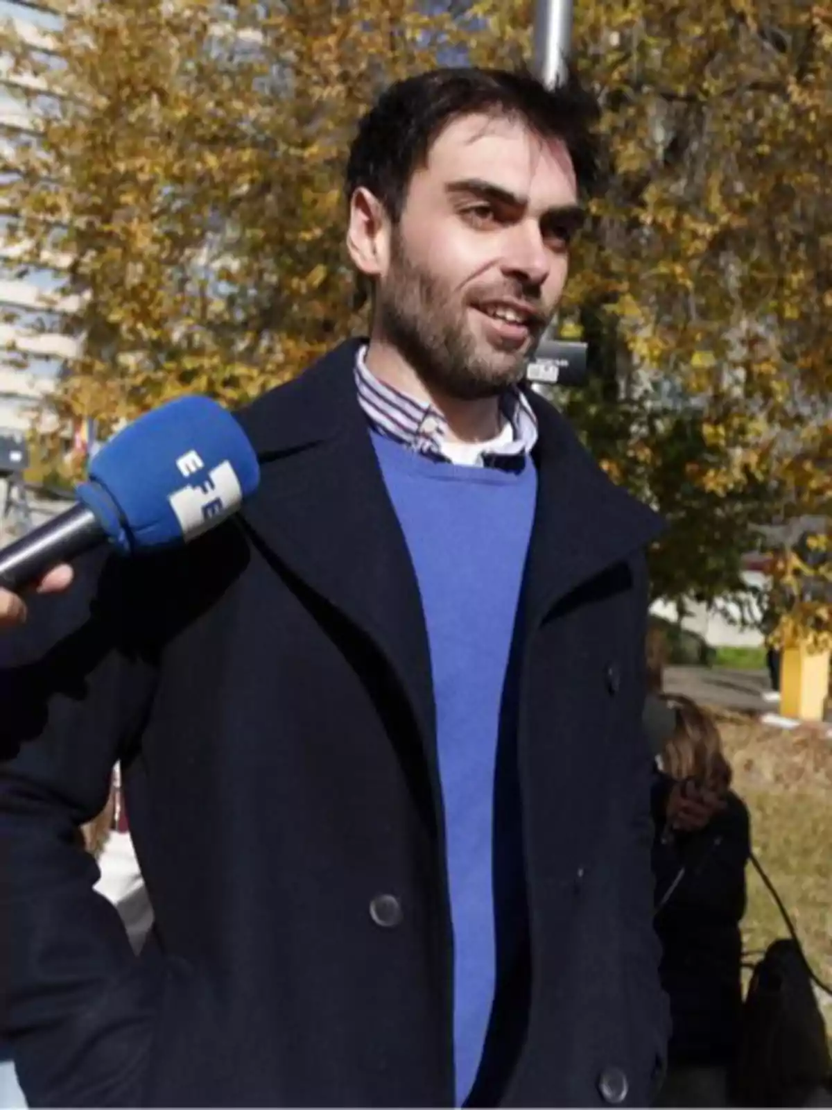 Hombre joven con barba y cabello oscuro vestido con abrigo azul y suéter azul claro siendo entrevistado al aire libre en un día soleado de otoño Hombre joven con barba y cabello oscuro vestido con abrigo azul y suéter azul claro siendo entrevistado al aire libre en un día soleado de otoño
