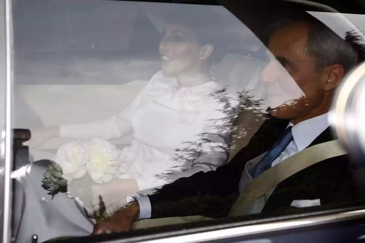 Una pareja vestida formalmente está sentada en el asiento trasero de un automóvil, con la mujer sosteniendo un ramo de flores blancas. Una pareja vestida formalmente está sentada en el asiento trasero de un automóvil, con la mujer sosteniendo un ramo de flores blancas.