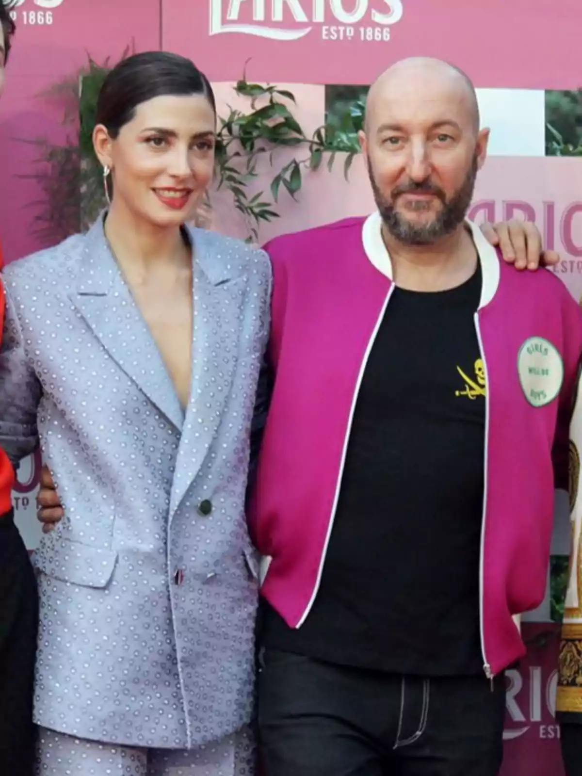 Dos personas posan juntas en un evento, una mujer con traje lila decorado y un hombre con chaqueta fucsia y camiseta negra, ambos sonríen frente a un fondo rosa con plantas. Dos personas posan juntas en un evento, una mujer con traje lila decorado y un hombre con chaqueta fucsia y camiseta negra, ambos sonríen frente a un fondo rosa con plantas.