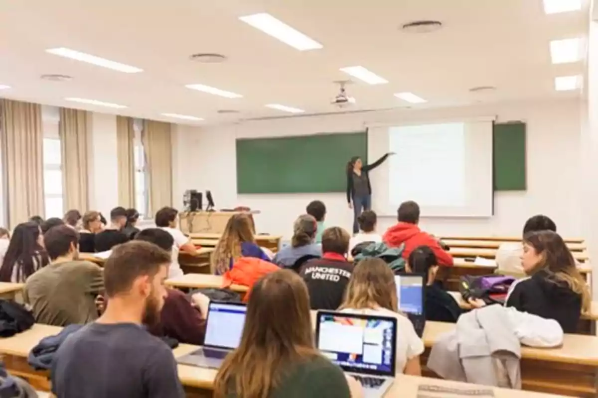 Estudiantes en un aula universitaria prestando atención a una profesora que está explicando en una pizarra.