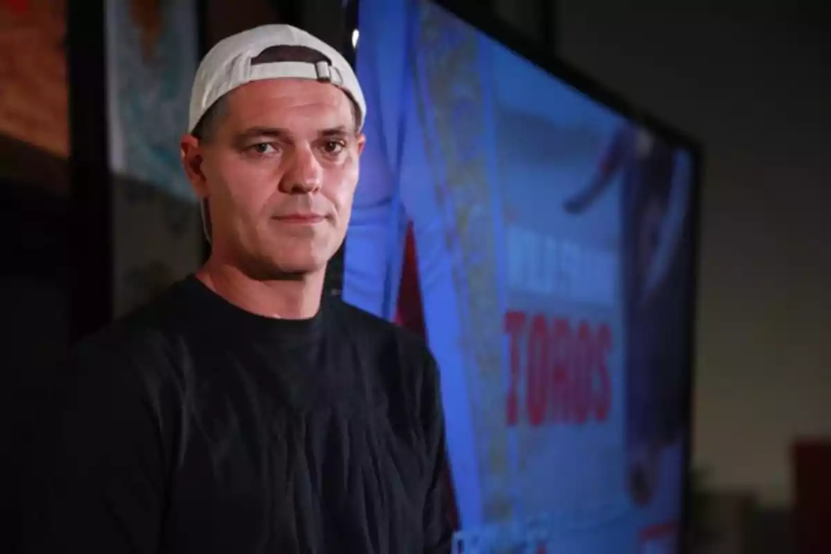 Un hombre con gorra blanca al revés y camiseta negra frente a una pantalla que muestra un cartel.