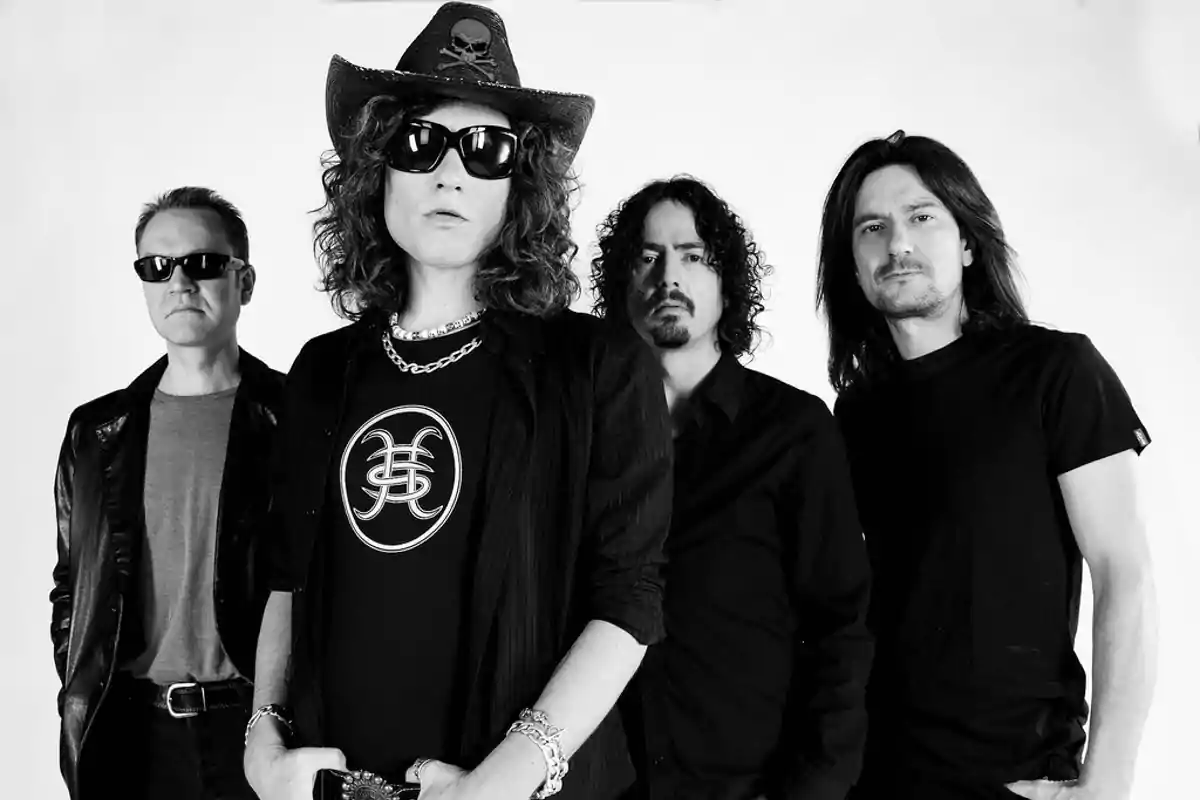 Cuatro hombres posan en blanco y negro, uno lleva sombrero y gafas de sol, todos visten de manera casual y seria.