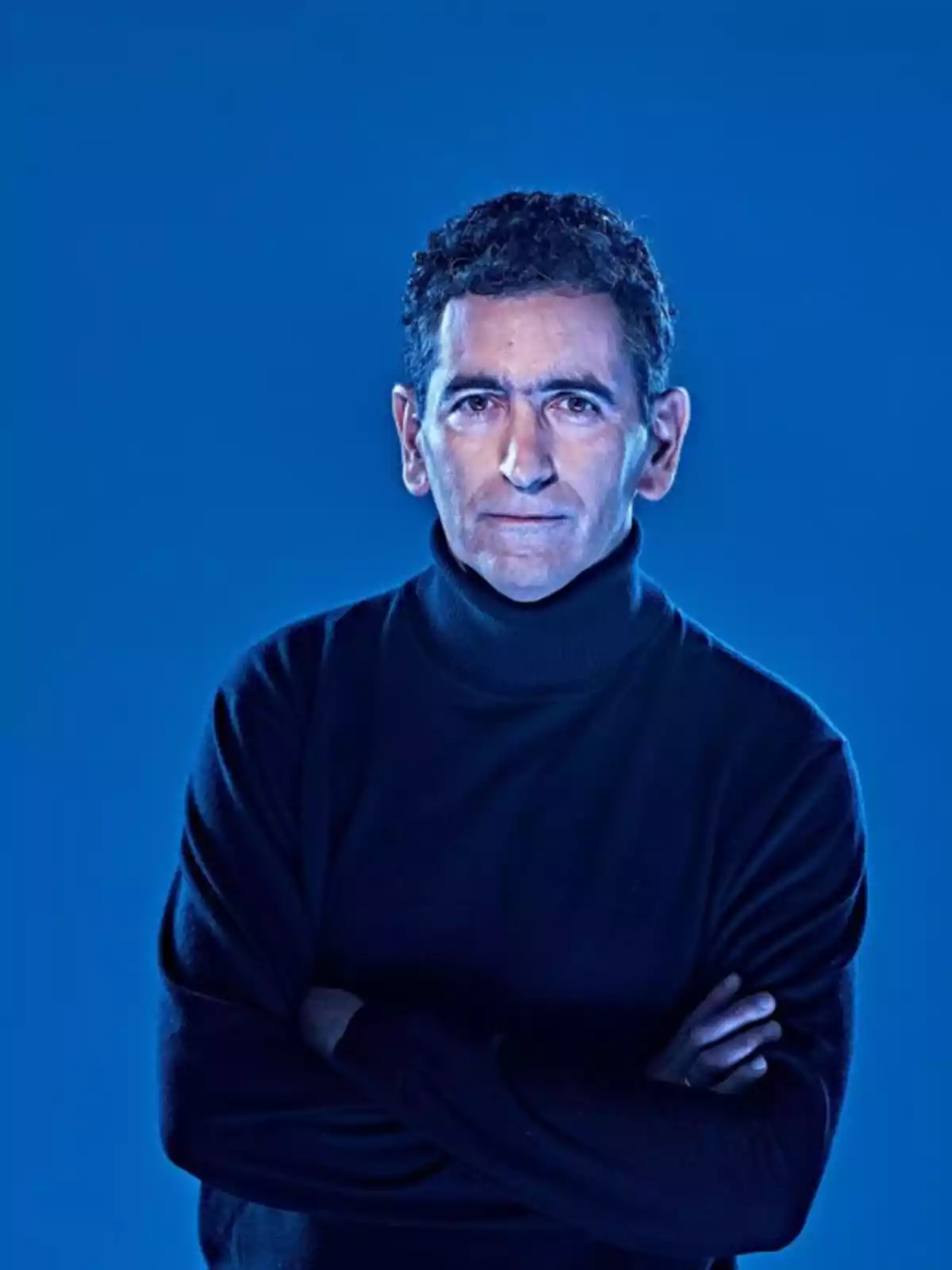 Hombre de cabello rizado y suéter negro posando con los brazos cruzados sobre un fondo azul.