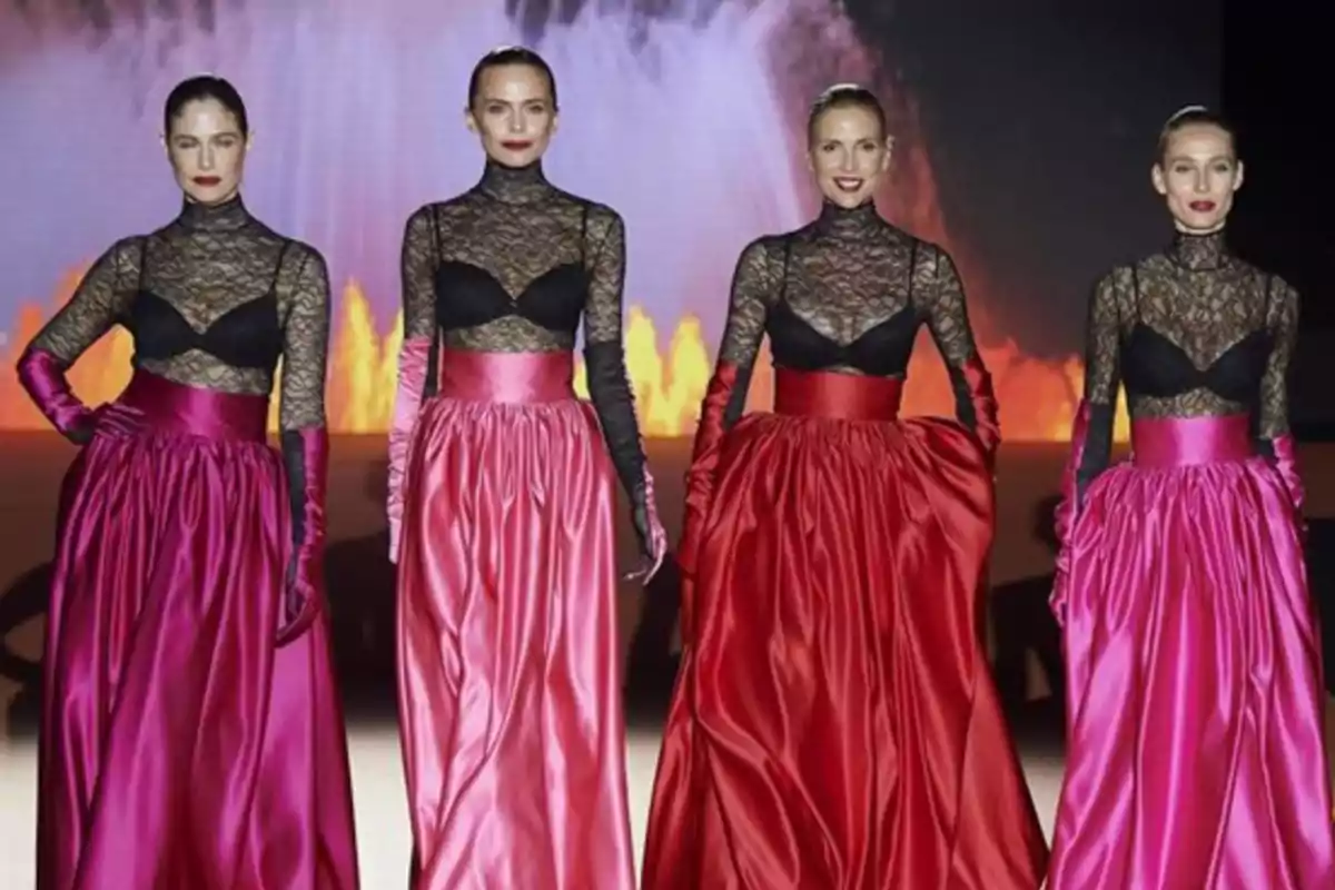 Cuatro modelos en la pasarela lucen conjuntos de encaje negro con faldas largas de satén en tonos fucsia y rojo, con un fondo de luces brillantes. Cuatro modelos en la pasarela lucen conjuntos de encaje negro con faldas largas de satén en tonos fucsia y rojo, con un fondo de luces brillantes.