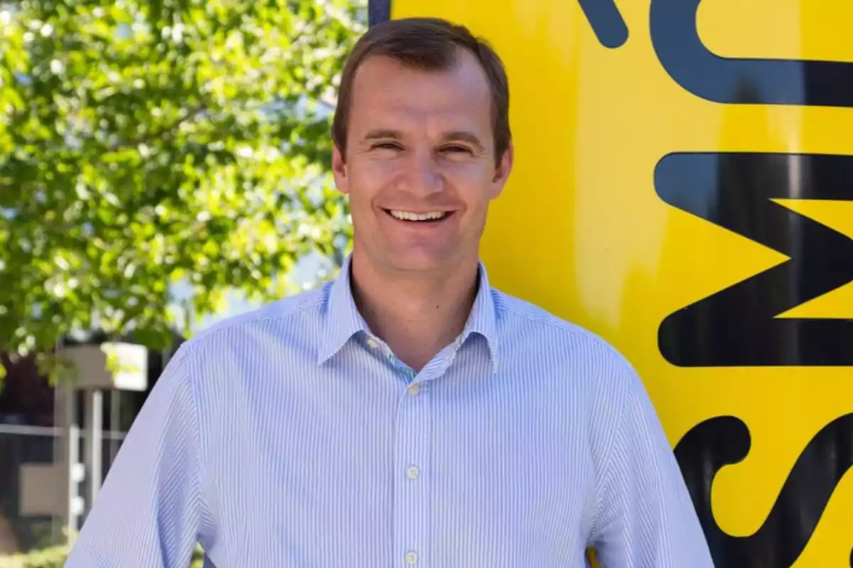 Hombre sonriente con camisa de rayas claras posando frente a un cartel amarillo y árboles verdes al fondo Hombre sonriente con camisa de rayas claras posando frente a un cartel amarillo y árboles verdes al fondo