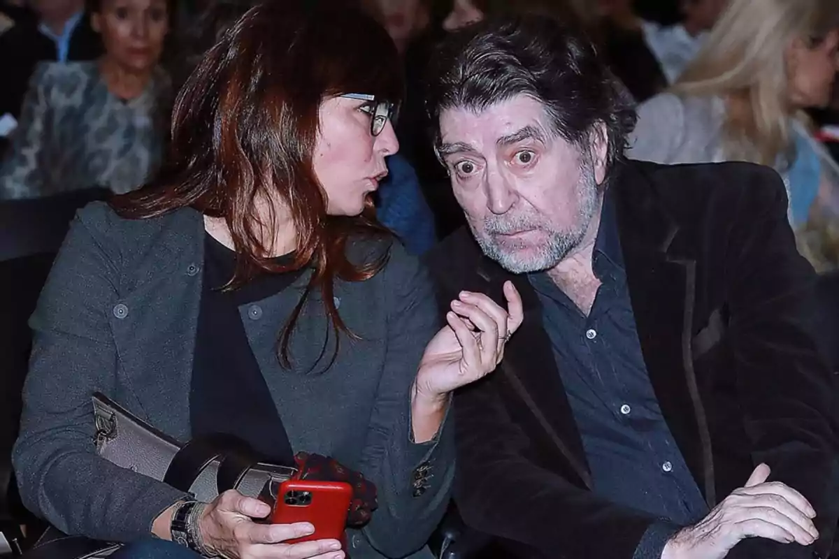 Una mujer conversa al oído con un hombre de cabello canoso y expresión sorprendida mientras están sentados en un evento rodeados de otras personas. Una mujer conversa al oído con un hombre de cabello canoso y expresión sorprendida mientras están sentados en un evento rodeados de otras personas.
