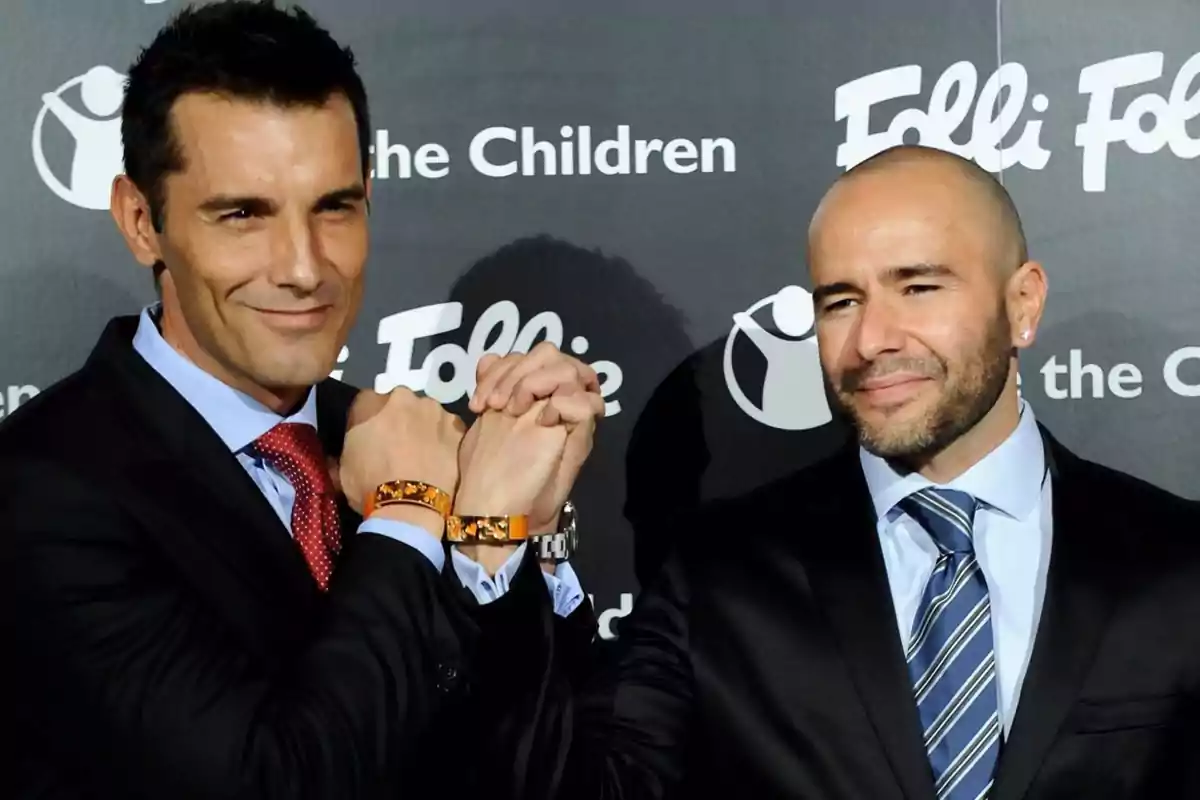Dos hombres vestidos con traje posan sonrientes en un evento, entrelazando sus manos y mostrando pulseras naranjas frente a un fondo con logotipos.