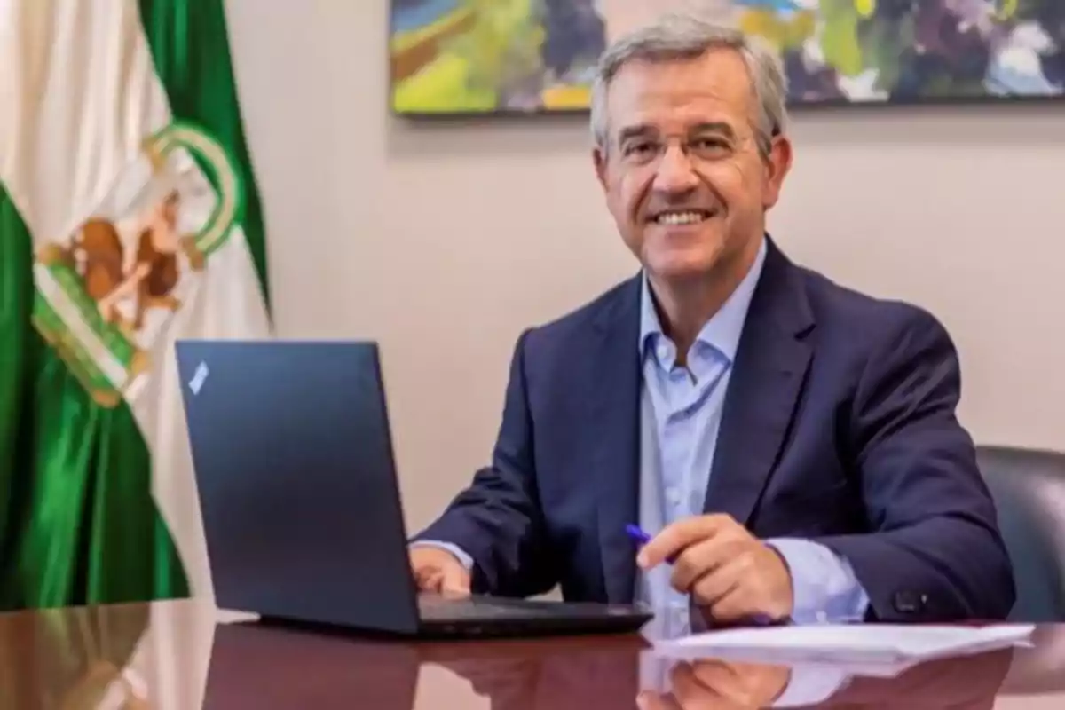 Hombre sonriendo mientras trabaja en una computadora portátil en una oficina con una bandera de fondo.