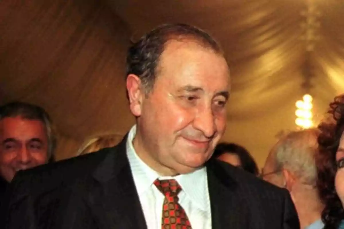 Un hombre mayor con traje y corbata sonríe en un evento social con varias personas alrededor. Un hombre mayor con traje y corbata sonríe en un evento social con varias personas alrededor.