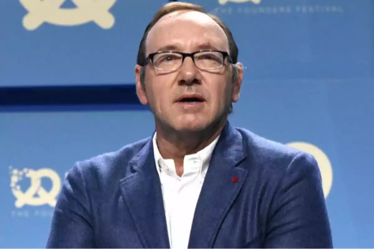 Un hombre con gafas y chaqueta azul está hablando frente a un fondo azul con logotipos. Un hombre con gafas y chaqueta azul está hablando frente a un fondo azul con logotipos.