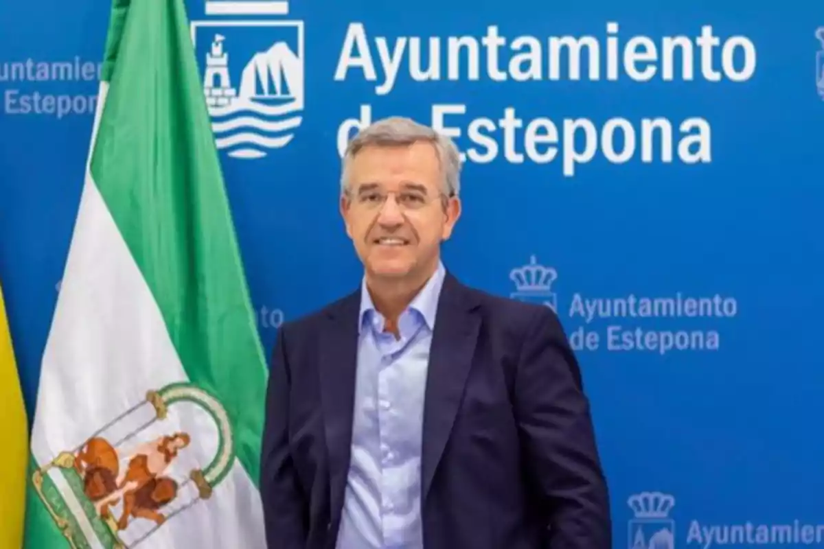 Hombre de pie frente a un fondo azul con el texto "Ayuntamiento de Estepona" y una bandera verde y blanca a su lado.