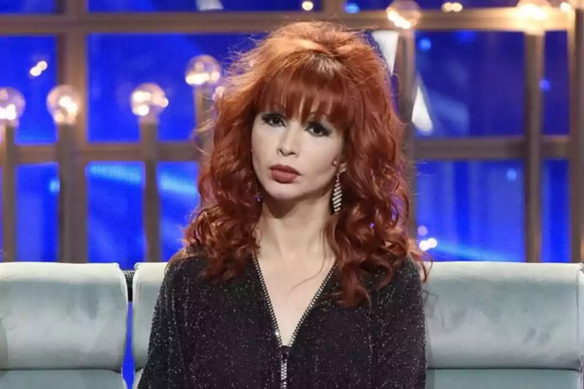 Mujer con cabello rojo rizado y vestido negro brillante sentada en un set de televisión iluminado.