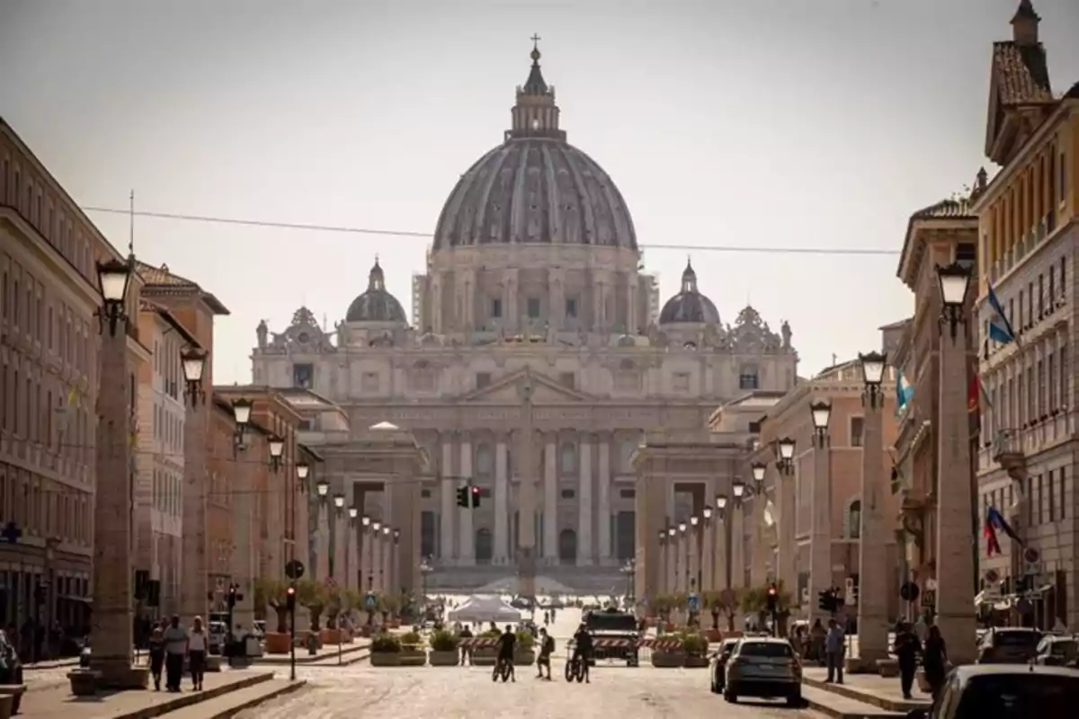 Vista frontal de la Basílica de San Pedro en el Vaticano con una calle flanqueada por edificios y farolas.