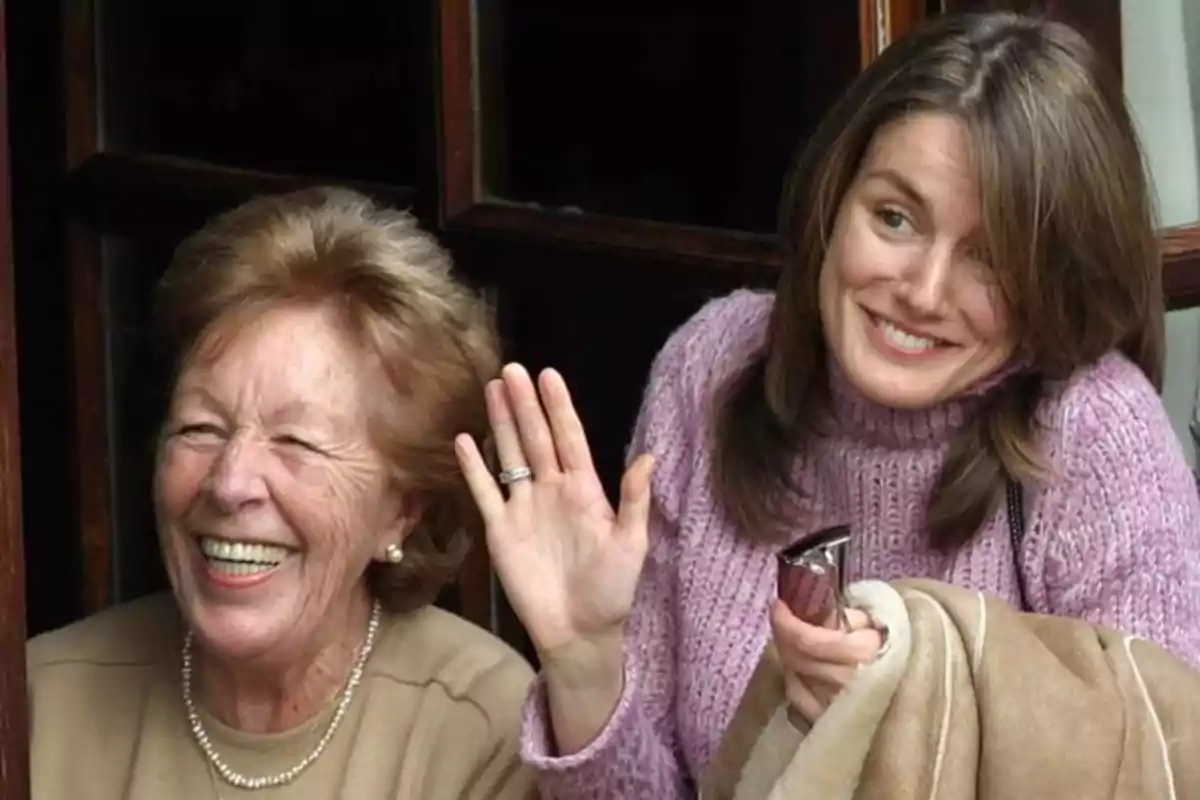 Dos mujeres sonrientes, una de ellas saludando con la mano, mientras están de pie cerca de una ventana. Dos mujeres sonrientes, una de ellas saludando con la mano, mientras están de pie cerca de una ventana.