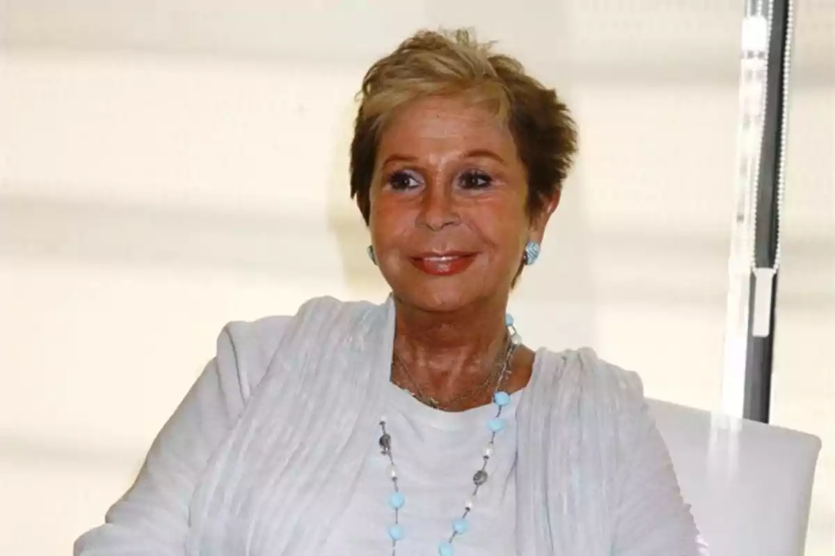 Una mujer sonriente con cabello corto y rubio vestida con una blusa blanca y un collar de cuentas azules y blancas.