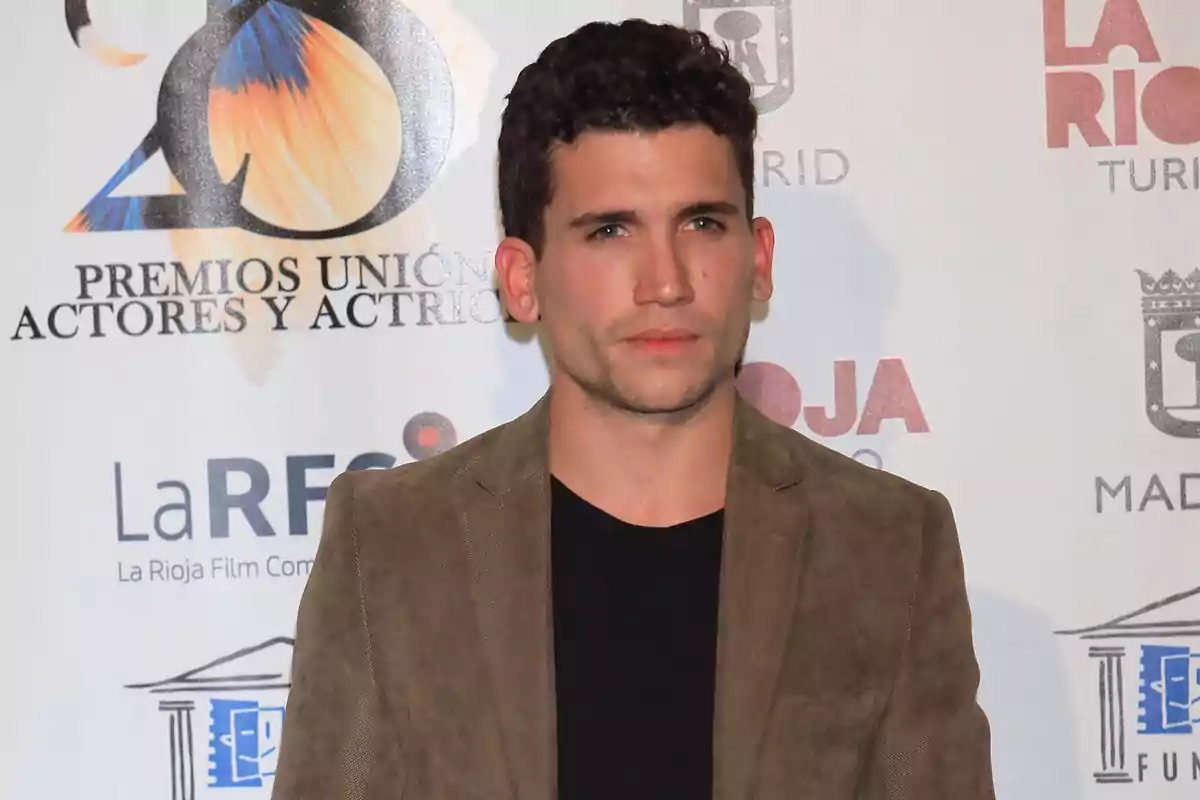 Hombre joven de cabello corto y rizado con chaqueta marrón y camiseta negra posando frente a un fondo con logotipos de premios y patrocinadores. Hombre joven de cabello corto y rizado con chaqueta marrón y camiseta negra posando frente a un fondo con logotipos de premios y patrocinadores.