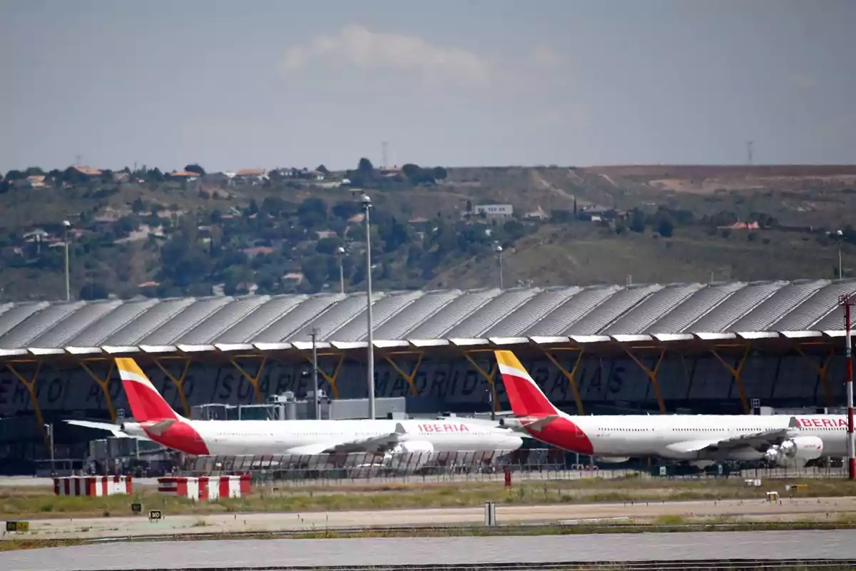 Aviones de la aerolínea Iberia estacionados frente a una terminal en un aeropuerto con colinas y casas al fondo.