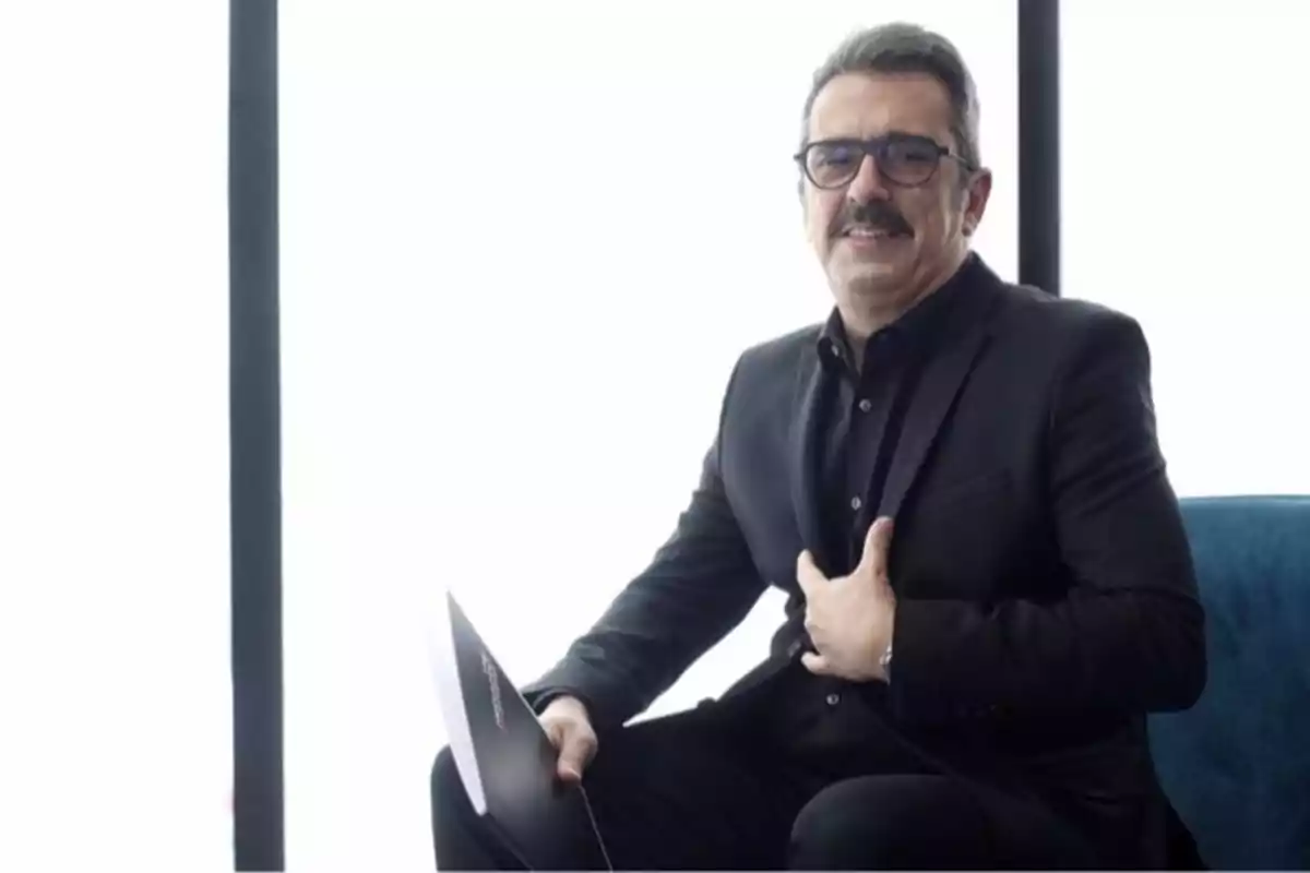 Un hombre con gafas y bigote, vestido con un traje negro, está sentado en una silla azul sosteniendo una carpeta. Un hombre con gafas y bigote, vestido con un traje negro, está sentado en una silla azul sosteniendo una carpeta.