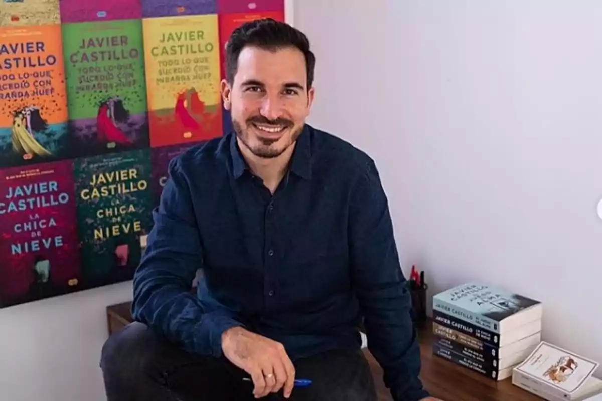 Un hombre sonriente sentado frente a una pared con portadas de libros y una pila de libros a su lado.