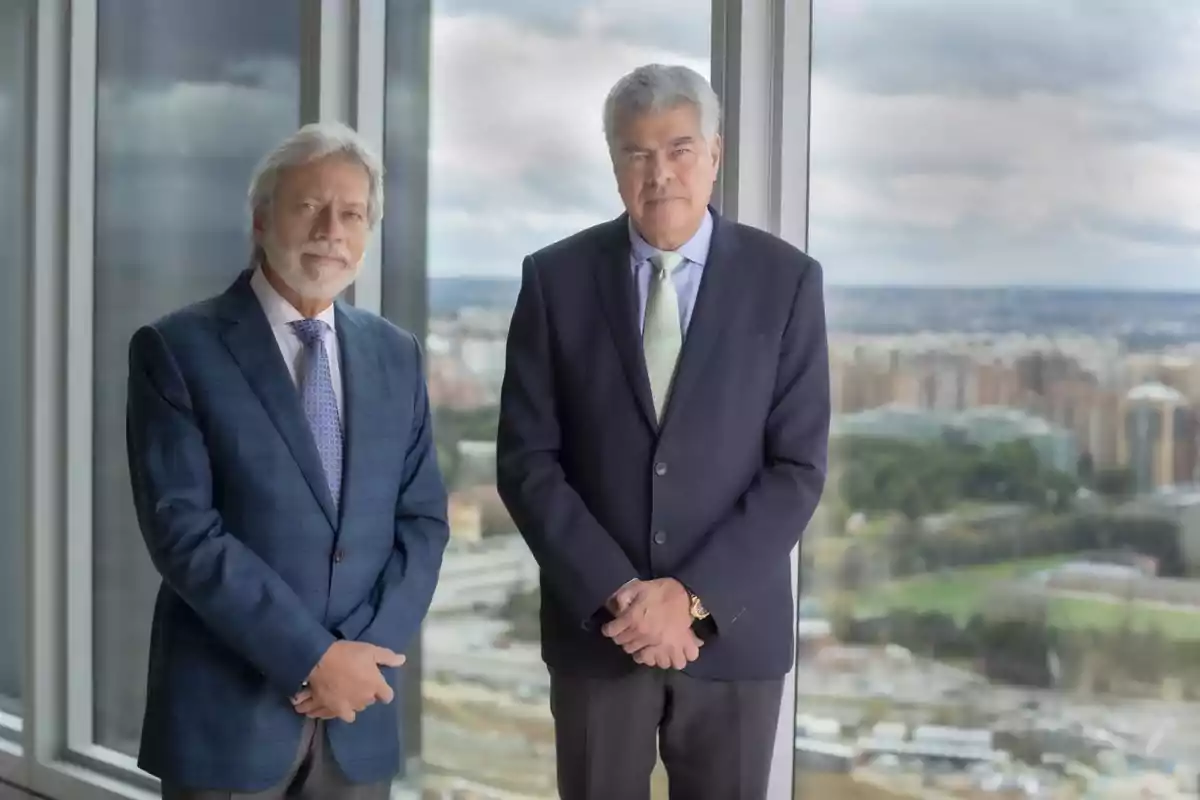 Dos hombres de negocios posan juntos frente a una ventana con vista a una ciudad.