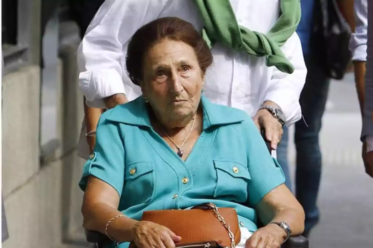 Mujer mayor con camisa turquesa sentada en una silla de ruedas mientras es empujada por otra persona en la calle Mujer mayor con camisa turquesa sentada en una silla de ruedas mientras es empujada por otra persona en la calle