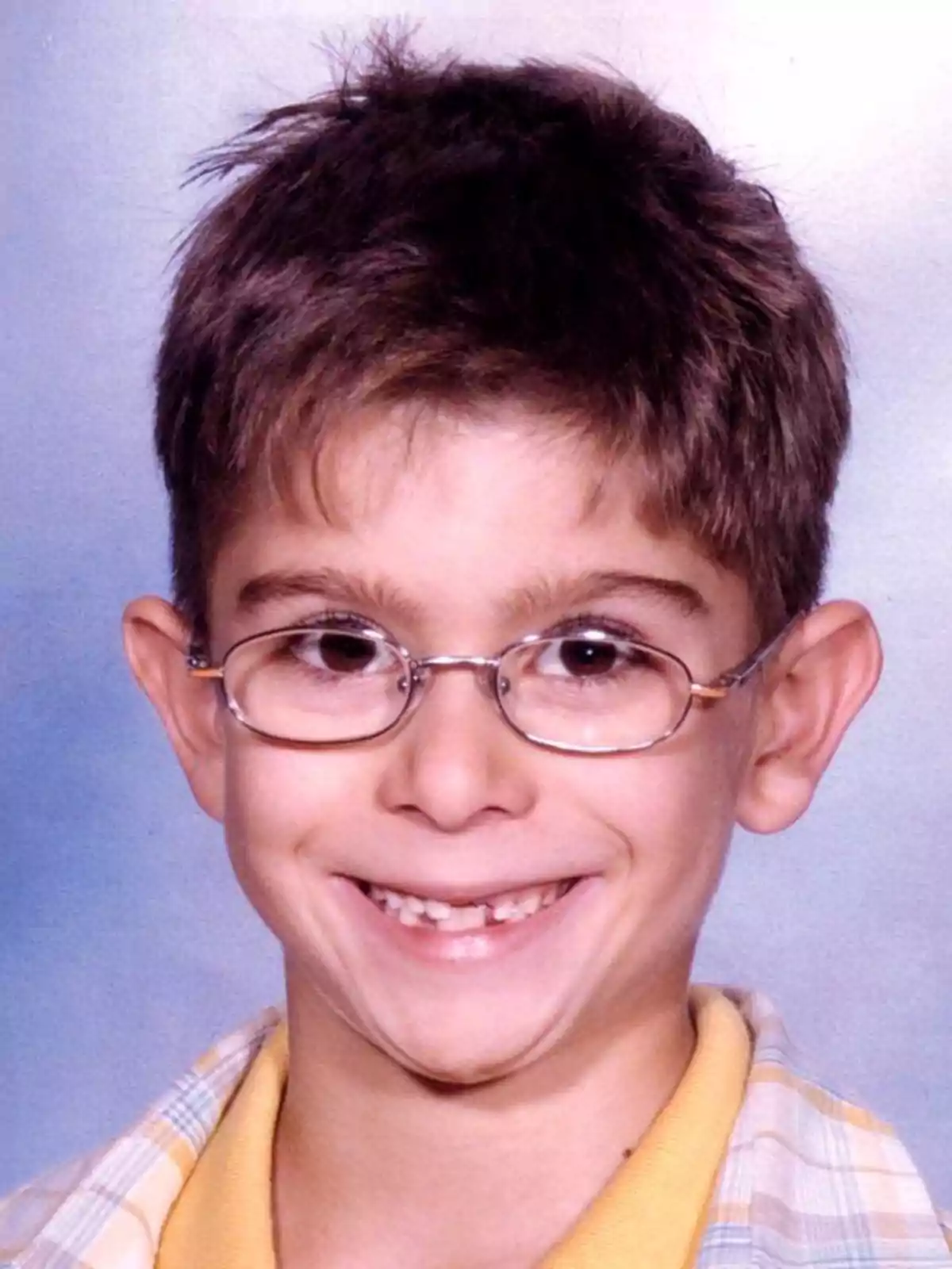 Un niño con gafas y una camisa a cuadros sonríe frente a un fondo azul.