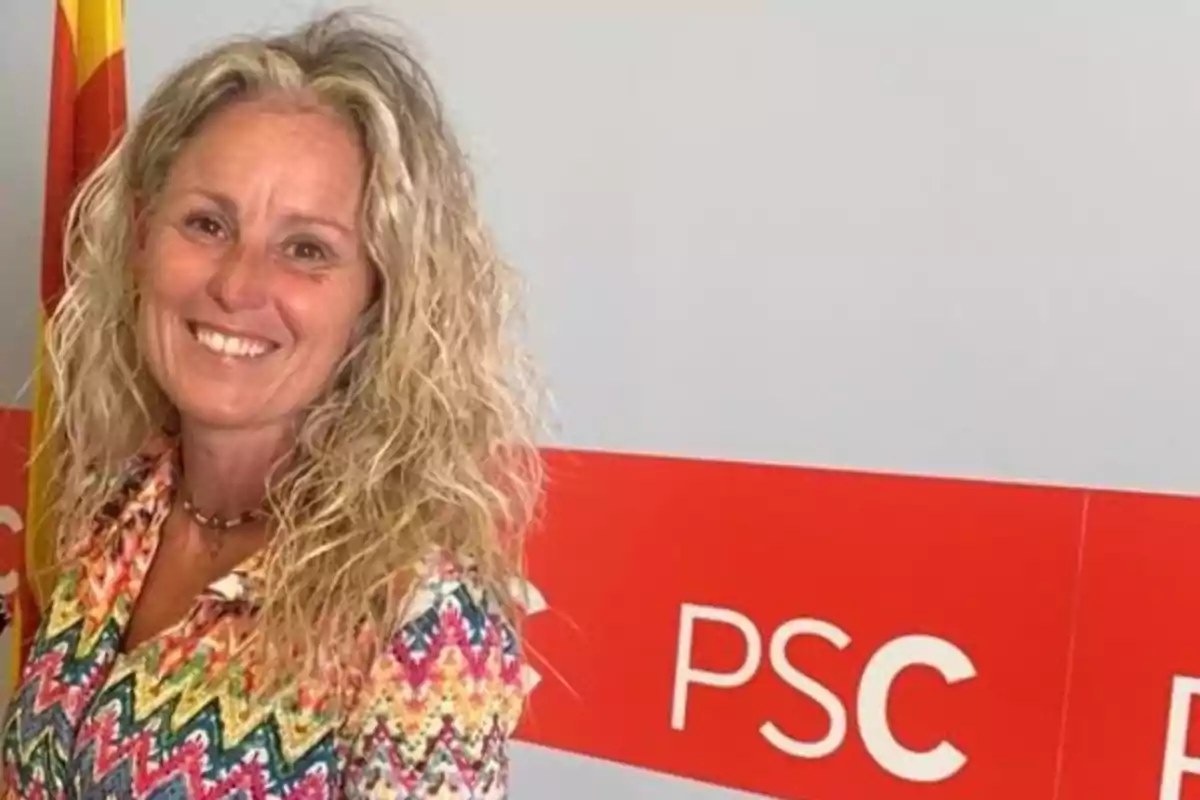 Mujer sonriente de cabello rubio rizado con camisa colorida posando frente a un fondo gris con un cartel rojo que tiene las letras PSC y una bandera a la izquierda Mujer sonriente de cabello rubio rizado con camisa colorida posando frente a un fondo gris con un cartel rojo que tiene las letras PSC y una bandera a la izquierda