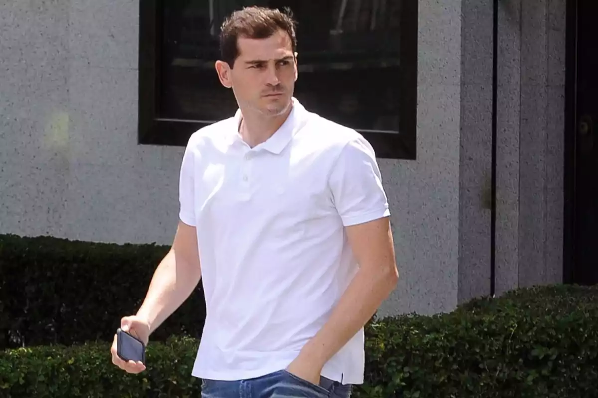 Hombre con camiseta polo blanca y pantalón de mezclilla sosteniendo un teléfono móvil mientras camina al aire libre Hombre con camiseta polo blanca y pantalón de mezclilla sosteniendo un teléfono móvil mientras camina al aire libre