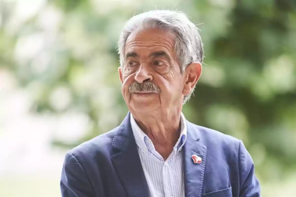 Un hombre mayor con bigote y cabello canoso viste una chaqueta azul y camisa a cuadros, mirando hacia un lado con un fondo desenfocado de vegetación.