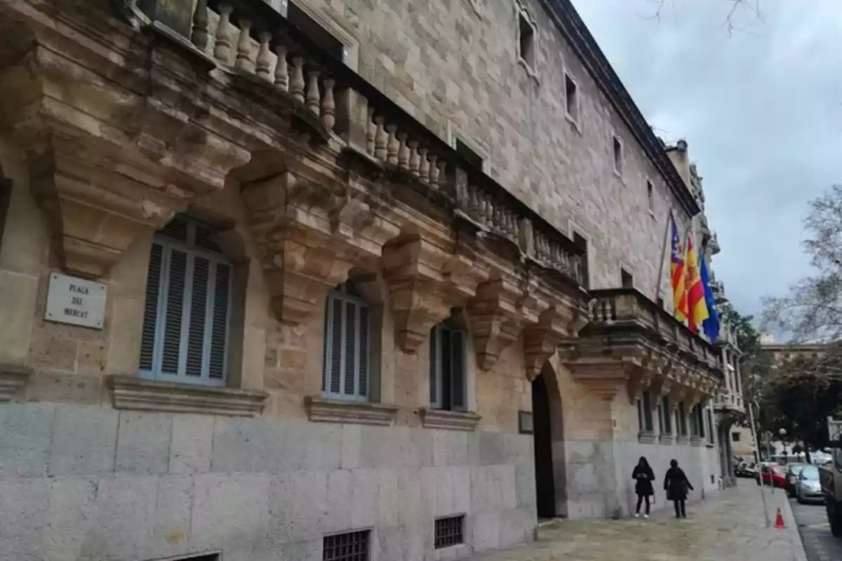 Fachada de un edificio histórico de piedra con balcones y banderas, dos personas caminando por la acera y cielo nublado Fachada de un edificio histórico de piedra con balcones y banderas, dos personas caminando por la acera y cielo nublado