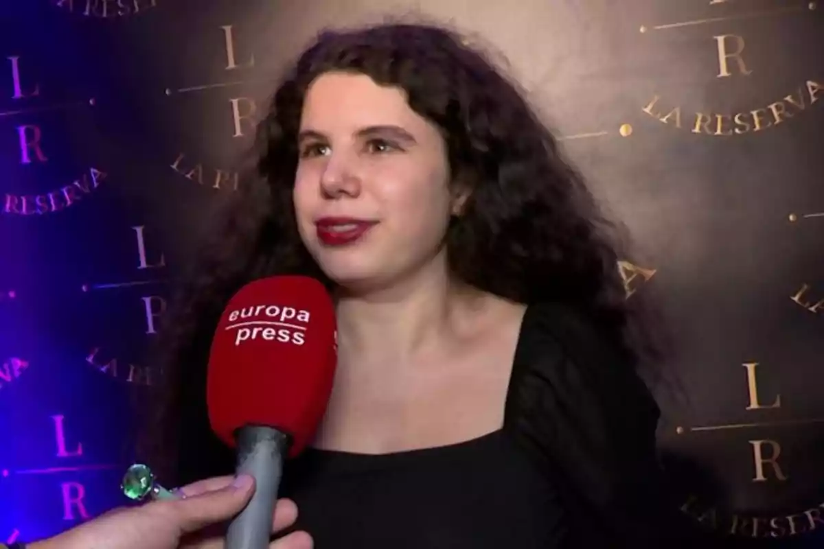 Una mujer con cabello rizado y largo está siendo entrevistada por un micrófono de Europa Press frente a un fondo con el logo de 