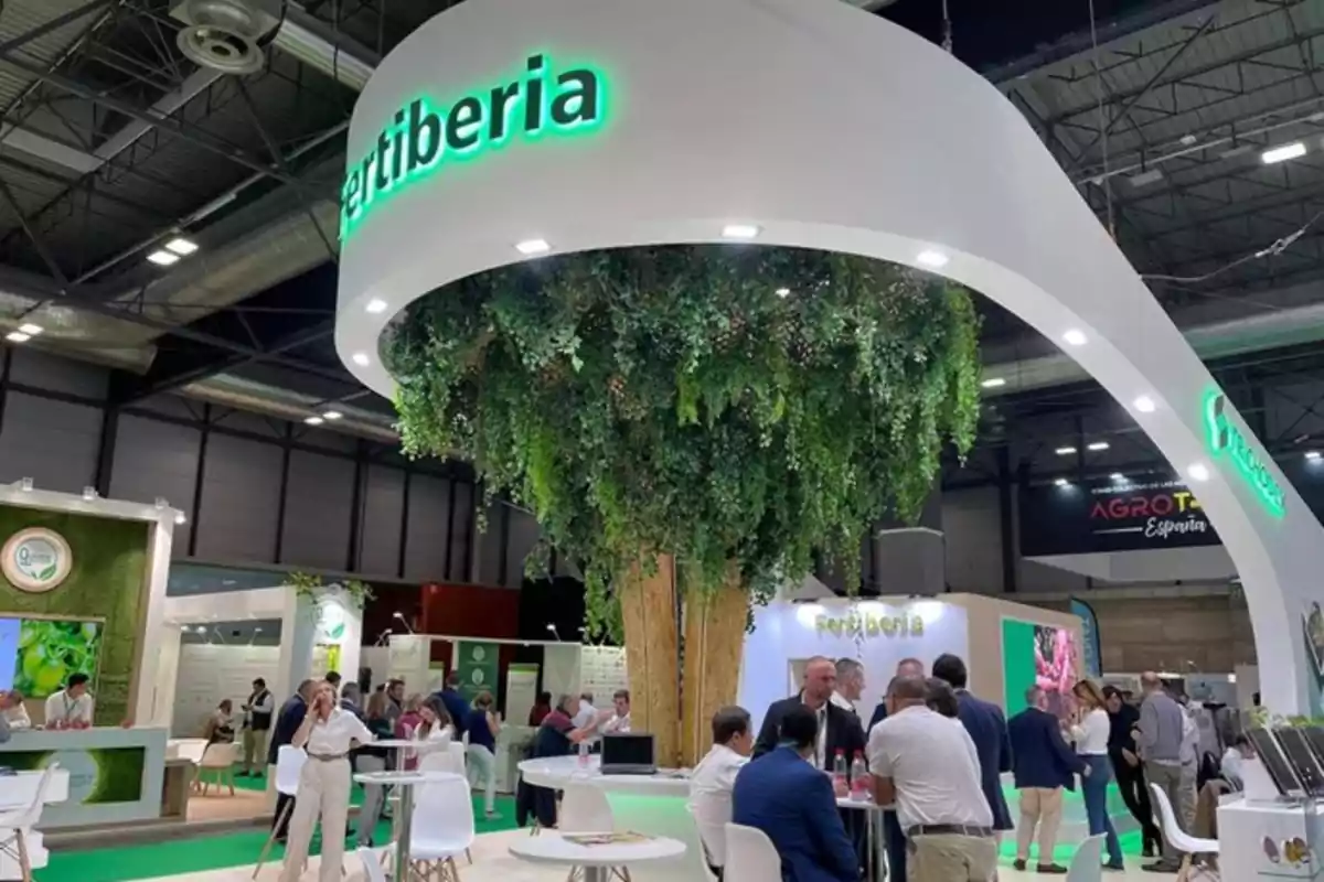 Personas interactuando en un stand de exhibición con un diseño moderno y vegetación colgante en un evento interior. Personas interactuando en un stand de exhibición con un diseño moderno y vegetación colgante en un evento interior.