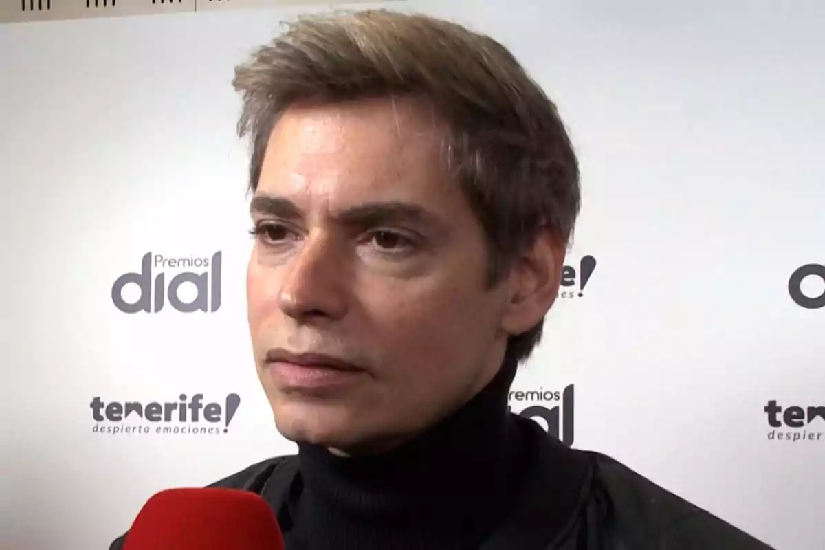 Un hombre con cabello claro y suéter oscuro sostiene un micrófono rojo frente a un fondo con logotipos de Un hombre con cabello claro y suéter oscuro sostiene un micrófono rojo frente a un fondo con logotipos de