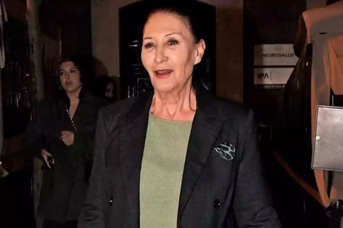Una mujer de cabello oscuro y recogido, vestida con un suéter verde y un abrigo oscuro, camina por un lugar con poca iluminación, mientras otra persona aparece en el fondo. Una mujer de cabello oscuro y recogido, vestida con un suéter verde y un abrigo oscuro, camina por un lugar con poca iluminación, mientras otra persona aparece en el fondo.