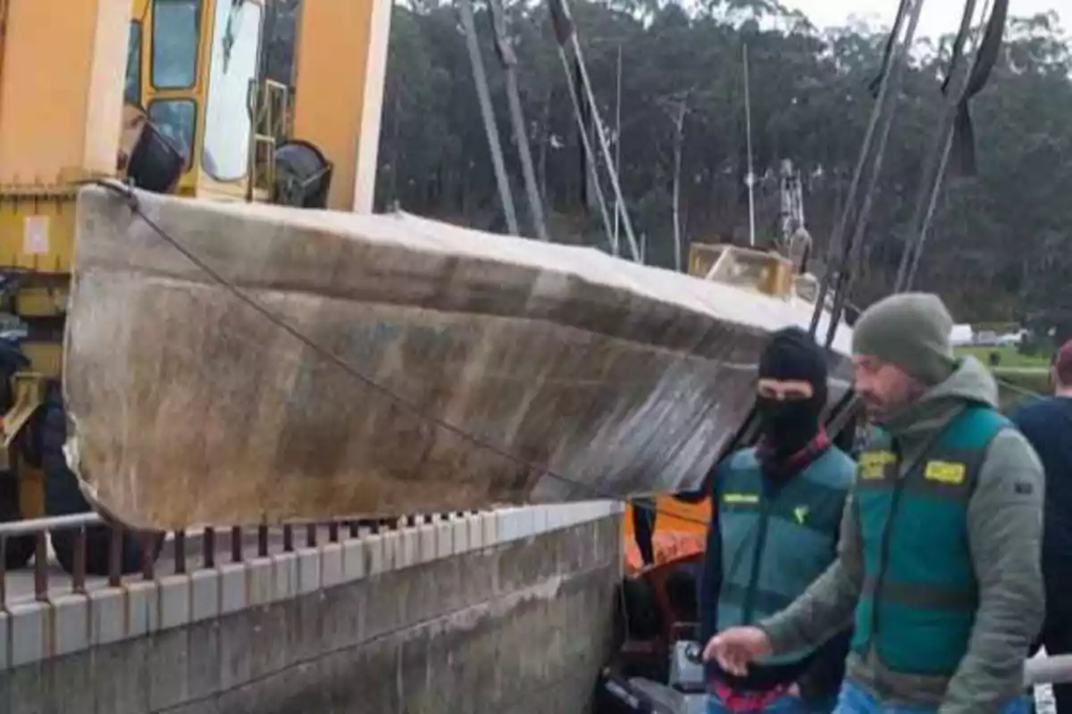 Un submarino de aspecto desgastado está siendo levantado por una grúa mientras dos personas con chalecos verdes observan la operación.