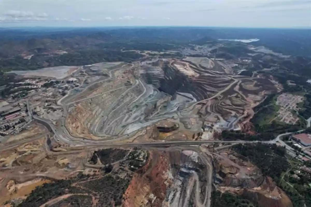 Vista aérea de una mina a cielo abierto con caminos serpenteantes y áreas de extracción rodeadas de vegetación y montañas en el horizonte.