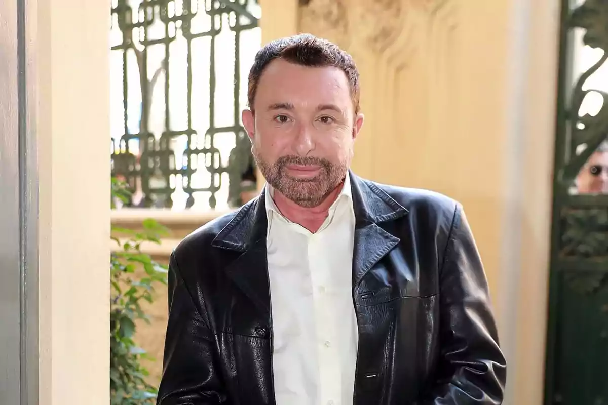 Hombre de cabello corto y barba con chaqueta de cuero negra y camisa blanca posando en un entorno elegante con fondo de rejas decorativas y plantas. Hombre de cabello corto y barba con chaqueta de cuero negra y camisa blanca posando en un entorno elegante con fondo de rejas decorativas y plantas.