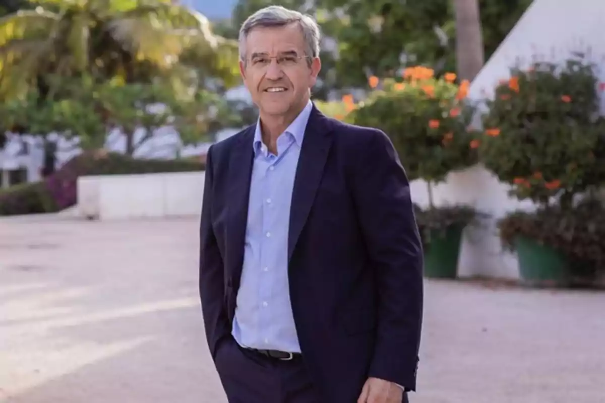 Hombre de pie al aire libre con traje oscuro y camisa azul, rodeado de vegetación y flores.