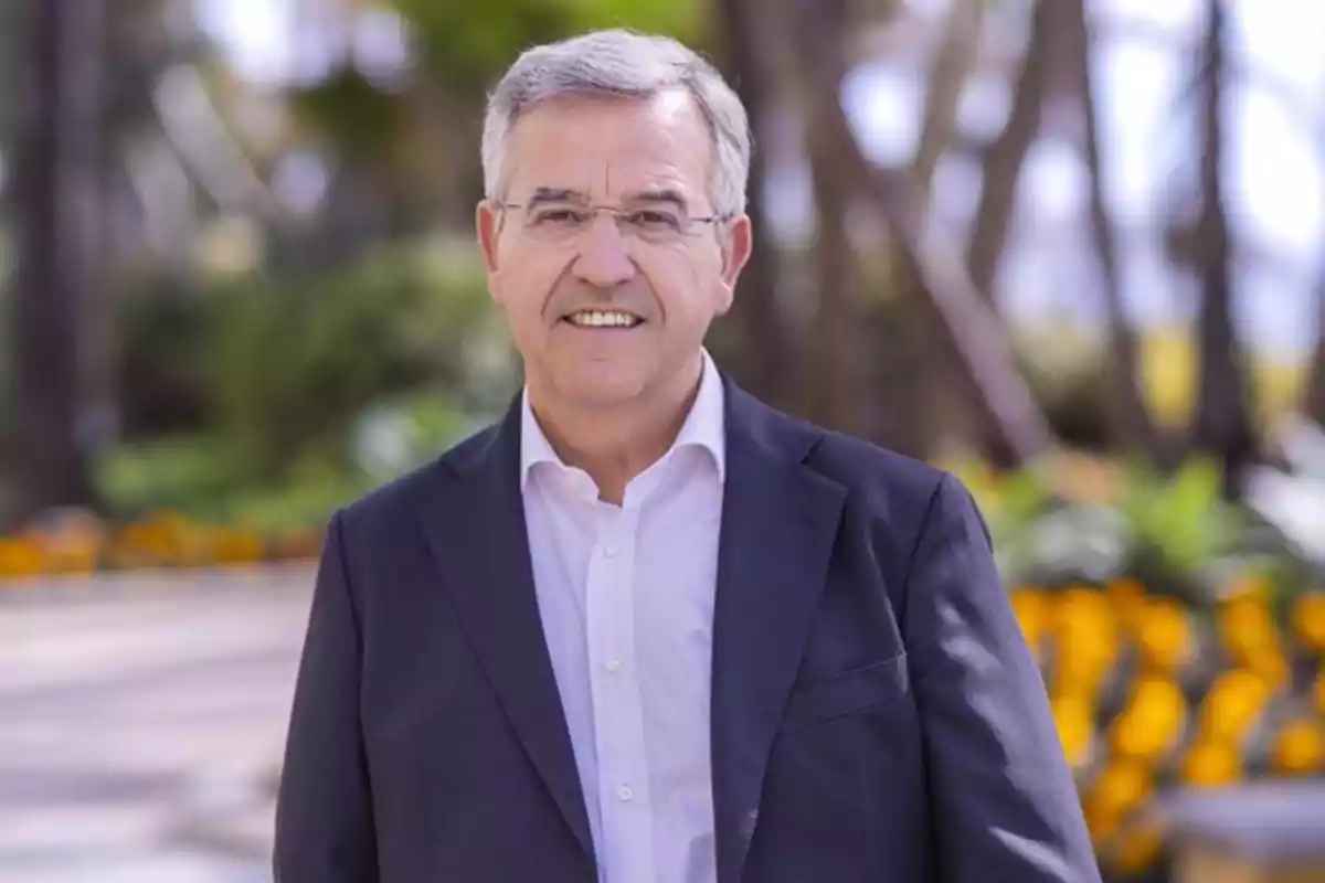 Un hombre mayor con cabello canoso y gafas sonríe mientras viste un traje oscuro y una camisa blanca en un entorno al aire libre con flores y árboles desenfocados al fondo.