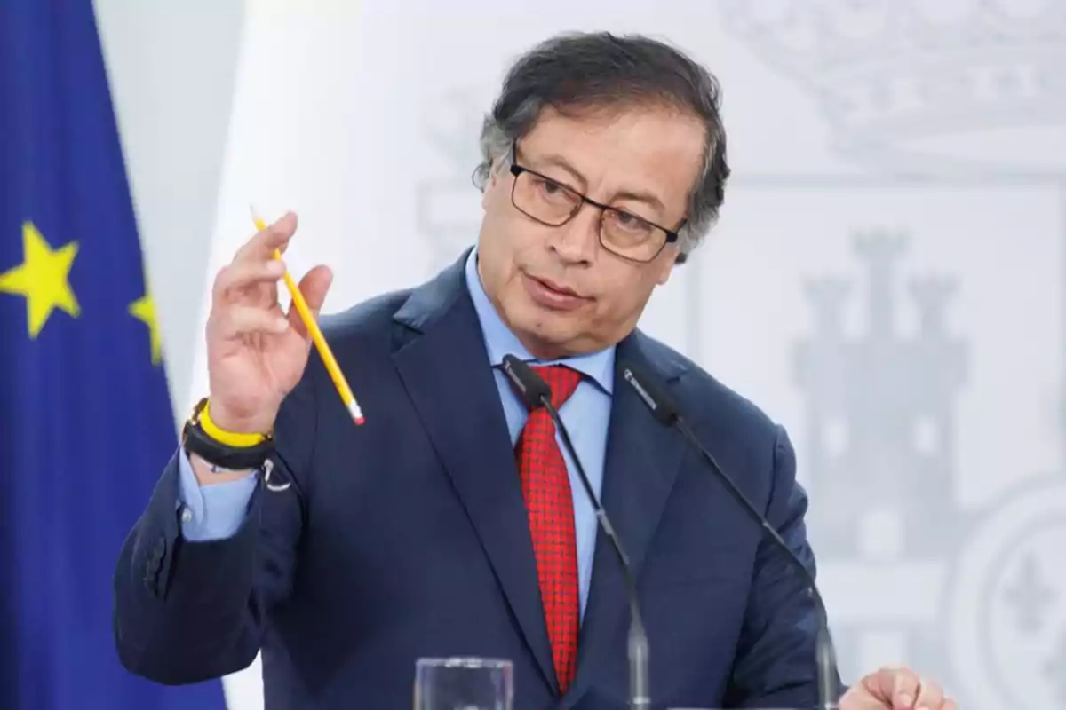 Hombre con gafas y traje azul hablando en una conferencia mientras sostiene un lápiz amarillo frente a dos micrófonos Hombre con gafas y traje azul hablando en una conferencia mientras sostiene un lápiz amarillo frente a dos micrófonos