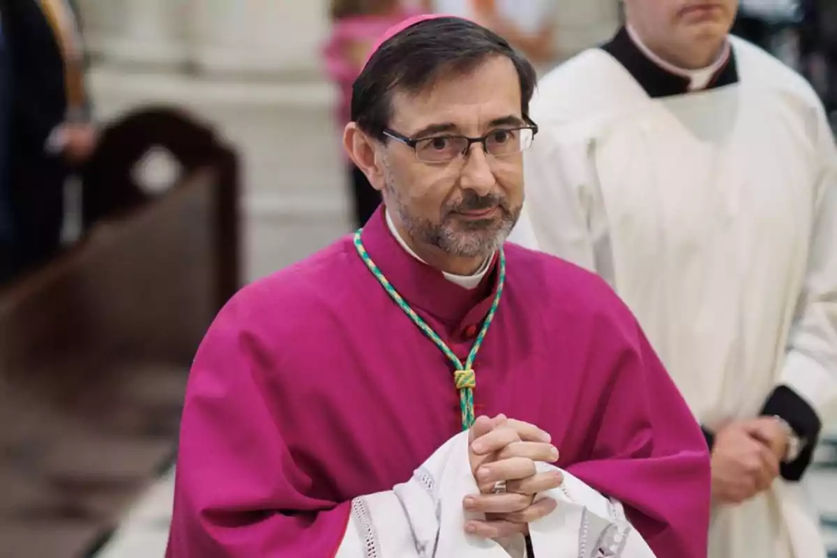 Un hombre con vestimenta religiosa de color púrpura y gafas está de pie con las manos juntas en un entorno eclesiástico.