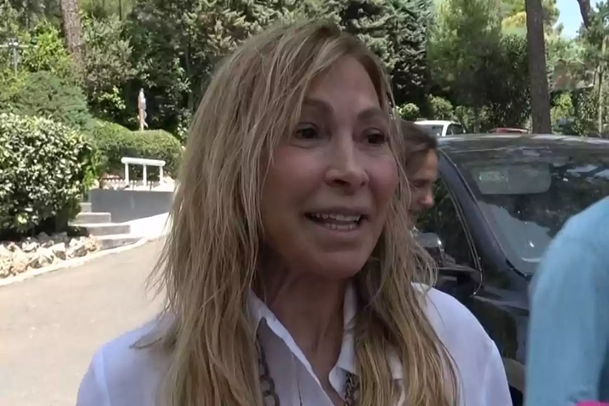 Una mujer sonriente con cabello rubio y largo en un entorno al aire libre con árboles y un coche al fondo.