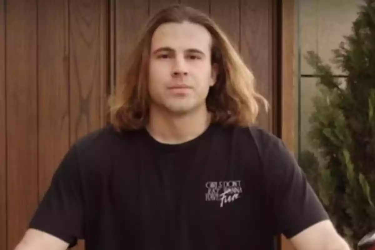 Un hombre con cabello largo y una camiseta negra está de pie frente a una puerta de madera.